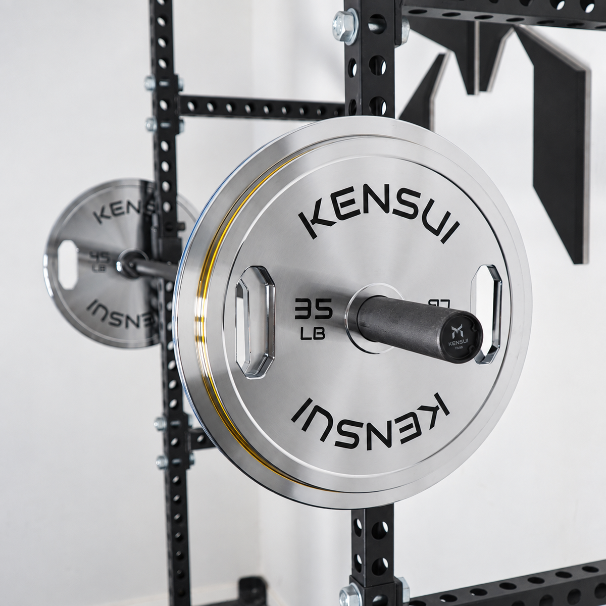 Kensui chrome grip calibrated plates on a barbell in a power rack, showing the 35 LB plate and yellow rim stripe.