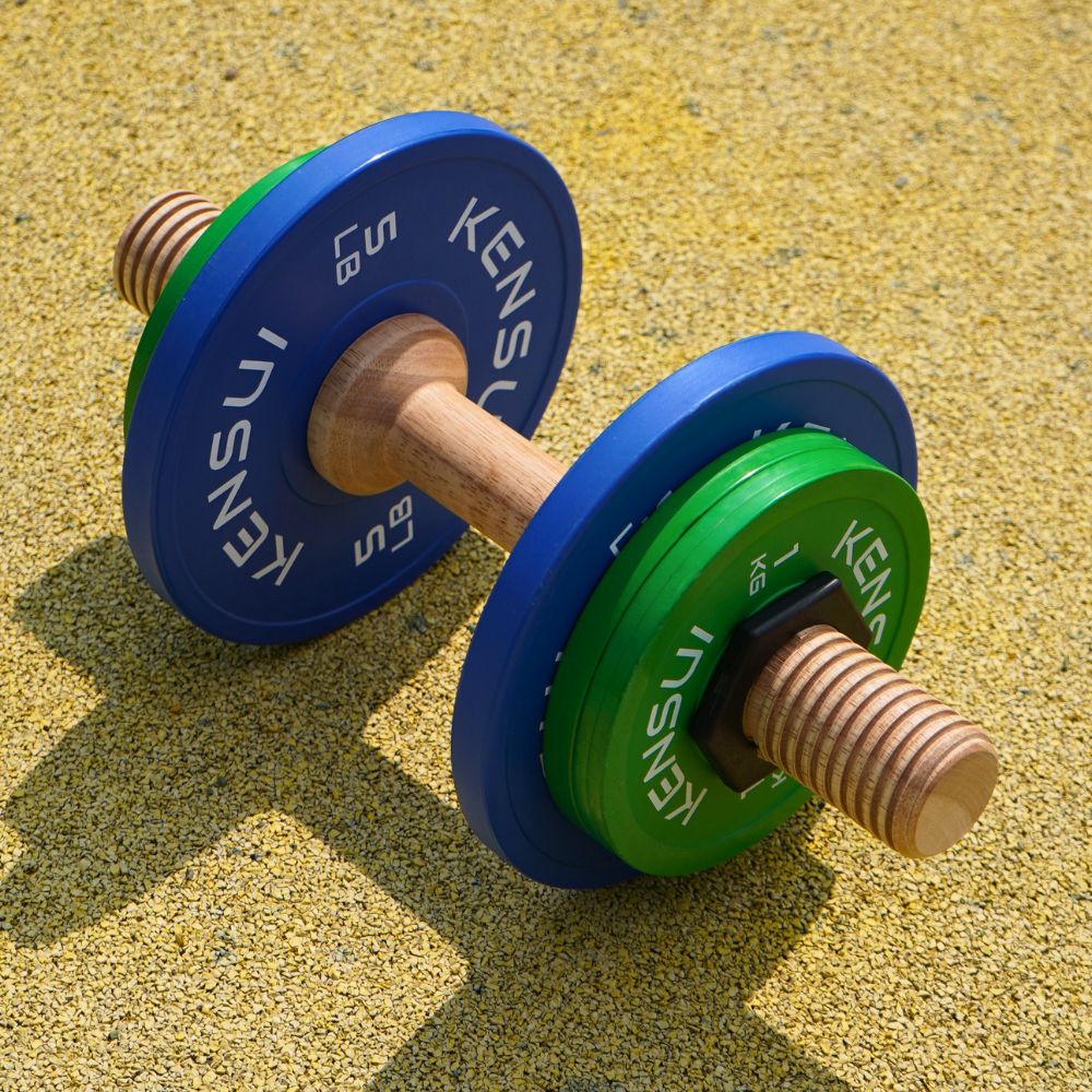 Timber wooden dumbbell prototype with blue and green plates on an outdoor training surface